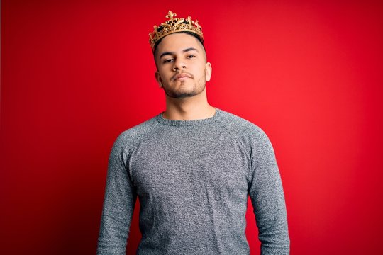 Young handsome man wearing golden crown of prince over isolated red background Relaxed with serious expression on face. Simple and natural looking at the camera.