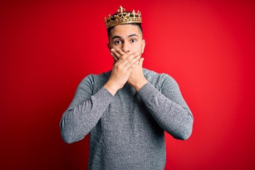 Young handsome man wearing golden crown of prince over isolated red background shocked covering mouth with hands for mistake. Secret concept.
