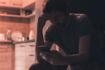 Depressed man typing text message on mobile phone in dark room