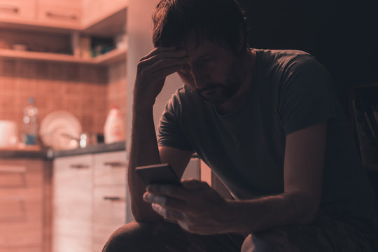 Depressed man typing text message on mobile phone in dark room