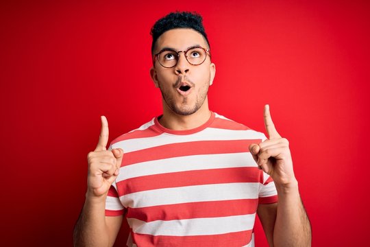 Young handsome man wearing casual striped t-shirt and glasses over isolated red background amazed and surprised looking up and pointing with fingers and raised arms.