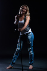 young, girl, blonde, jeans, clothes on a dark background emotionally sings into a microphone