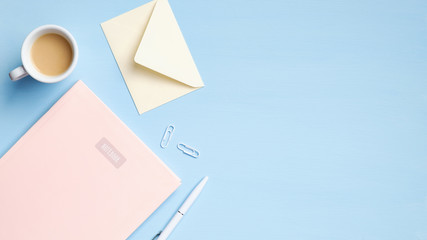 Flat lay, top view office table desk. Minimal style feminine workspace with pink notebook, cup of coffee, envelope, pen on blue background