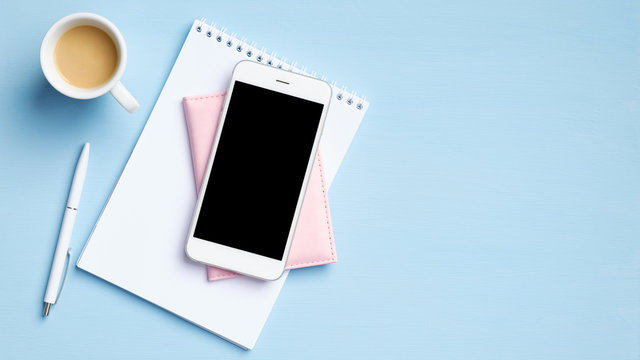 Modern office desk with smartphone with blank mockup screen, cup of coffee, blank paper notepad, document on blue background. Top view with copy space, flat lay