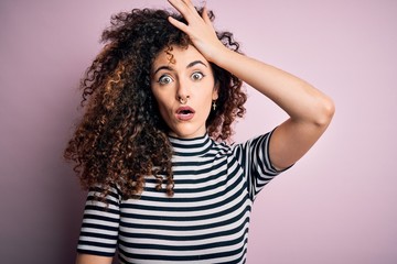 Young beautiful woman with curly hair and piercing wearing casual striped t-shirt surprised with hand on head for mistake, remember error. Forgot, bad memory concept.
