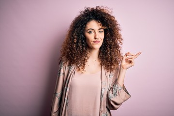 Young beautiful woman with curly hair and piercing wearing casual pink t-shirt with a big smile on face, pointing with hand and finger to the side looking at the camera.