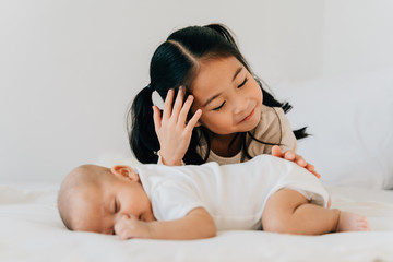 Asian family of cute little sister touching newborn baby boy brother. Toddler kid and new sibling relax in a white bedroom at home with love and tenderness
