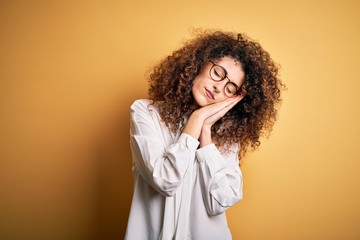 Young beautiful brunette woman with curly hair and piercing wearing shirt and glasses sleeping tired dreaming and posing with hands together while smiling with closed eyes.