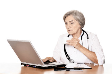 Serious elderly woman doctor sitting at table with computer on white