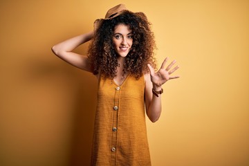 Beautiful brunette woman on vacation with curly hair and piercing wearing hat and dress showing and pointing up with fingers number five while smiling confident and happy.