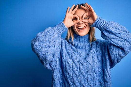 Young beautiful blonde woman wearing winter wool sweater over blue isolated background doing ok gesture like binoculars sticking tongue out, eyes looking through fingers. Crazy expression.