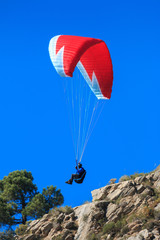 Paraglider in low flight over a rock and a tree during a sunny day.