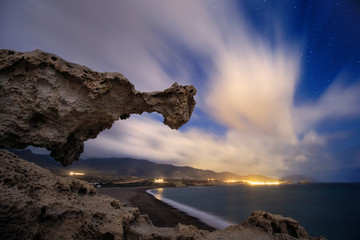 Night photography of long exposure, with rock formation in the foreground at Los Escullos beach and in the background the lights of La Isleta del Moro, Cabo de Gata Natural Park, Almería, Andalusia.