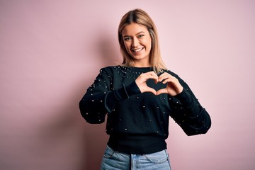 Young beautiful blonde woman wearing fashion sweater over pink isolated background smiling in love showing heart symbol and shape with hands. Romantic concept.