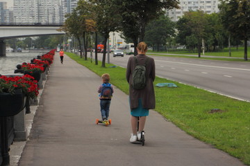 A woman and a child boy riding a kick scooters on a road in the Park at summer evening, outdoor vacation in big city