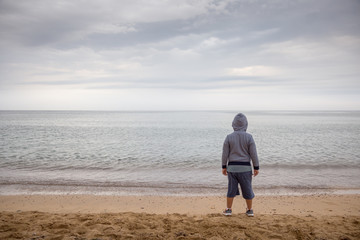Lonely hooded boy turned on his back looking out to sea on a gray and cloudy day.