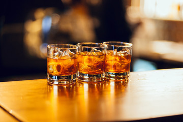 three glasses with whiskey and ice cubes on the bar with bokeh on background.