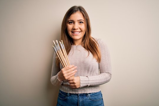 Young painter artist woman painting using paintbrush over isolated background with a happy face standing and smiling with a confident smile showing teeth