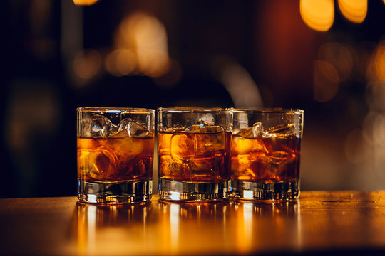 three glasses with whiskey and ice cubes on the bar with bokeh on background.