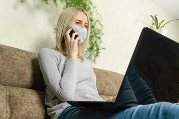 Young blonde woman in a medical mask with a laptop on her lap and a mobile phone in her hands is sitting on the sofa. Concept of work at home, self-isolation, pandemics.