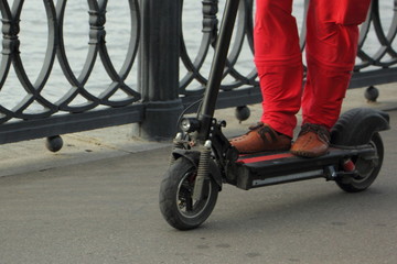 A man in red trousers ride on folding electrical kick scooter on asphalt road in Park on summer day, knee-deep front view close up, alternative eco transport