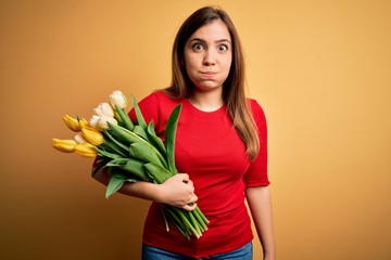 Young blonde woman holding romantic bouquet of tulips flowers over yellow background puffing cheeks with funny face. Mouth inflated with air, crazy expression.