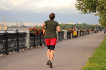 Street sport, one running mature woman in sportswear jogging along the fence of embankment alley of Moscow river on a summer evening, back view in Park in perspective