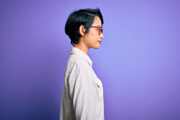 Young beautiful asian girl wearing casual shirt and glasses standing over purple background looking to side, relax profile pose with natural face and confident smile.