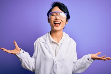 Young beautiful asian scientist girl wearing coat and glasses over purple background celebrating mad and crazy for success with arms raised and closed eyes screaming excited. Winner concept