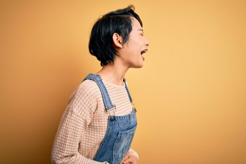 Young beautiful asian girl wearing casual denim overalls over isolated yellow background with hand on stomach because nausea, painful disease feeling unwell. Ache concept.