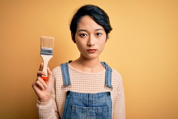 Young beautiful chinese woman painting holding paint brush over isolated yellow background with a confident expression on smart face thinking serious