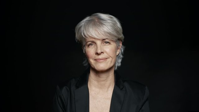 Close Up Of A Smiling Senior Woman With Short White Hair Looking At Camera. Close Up Of Happy Mature Woman In Formal Wear On Black Background.
