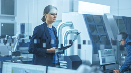 
Modern Factory: Confident Female Engineer Walks Through Facility, Holding Laptop, Inspecting and Monitoring Workshop Workings. Manufactory Has Professionals Working on CNC Machinery, Robot Arm