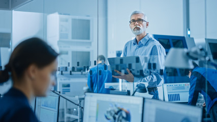 Modern Factory: Senior Engineer Looks at Camera, Holding Laptop, Inspecting, Overlooking Workshop...