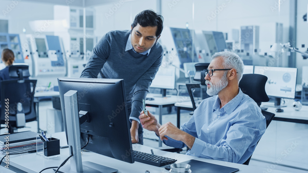 Poster Modern Factory Office: Indian Project Manager Talks to Caucasian Senior Engineer whos Working on Computer, Inspect Metal Component. Professionals Solving Heavy Industry Problems, Having Discussion 
