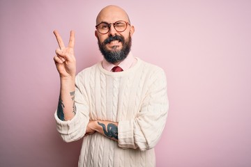 Handsome bald man with beard and tattoo wearing glasses and sweater over pink background smiling with happy face winking at the camera doing victory sign. Number two.