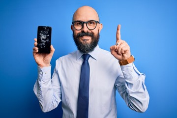 Handsome bald business man with beard holding broken smartphone showing cracked screen surprised with an idea or question pointing finger with happy face, number one