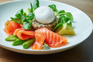 Tasty and healthy continental breakfast - quinoa with benedict eggs, spinach, avocado, salted salmon and asparagus in white bowl. Wooden background