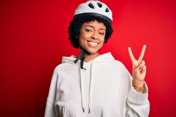 Young African American afro cyciling woman with curly hair wearing bike security helmet smiling with happy face winking at the camera doing victory sign. Number two.