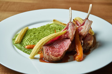 Rack of medium rare lamb with a side dish of green puree and baby corn and carrots on a white plate on a wooden background. Close up