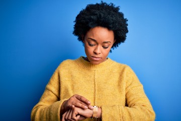 Young beautiful African American afro woman with curly hair wearing yellow casual sweater Checking the time on wrist watch, relaxed and confident