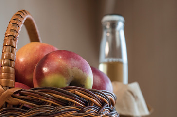 Basket with red fresh apples and a bottle of cider on a wooden table.