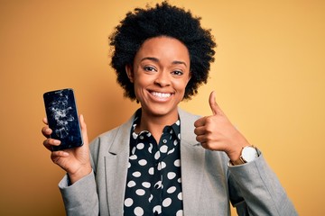 Young African American afro woman with curly hair holding cracked broken smartphone happy with big smile doing ok sign, thumb up with fingers, excellent sign