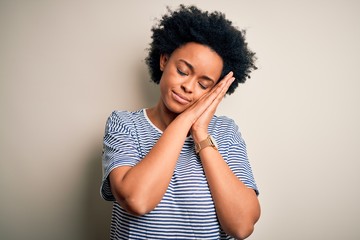 Young beautiful African American afro woman with curly hair wearing striped t-shirt sleeping tired dreaming and posing with hands together while smiling with closed eyes.