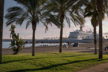 Beach panorama at sunset with palm trees in the grass near the sandy beach with straw umbrellas and cruise ship in Malaga Beach La Malagueta, Spain.