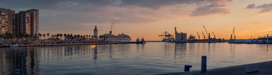 Sunset in Malaga Port wide panorama with purple cloudy sky. View of Quay 1 with palm trees and promenade, with cranes and a cruise ship sailing.