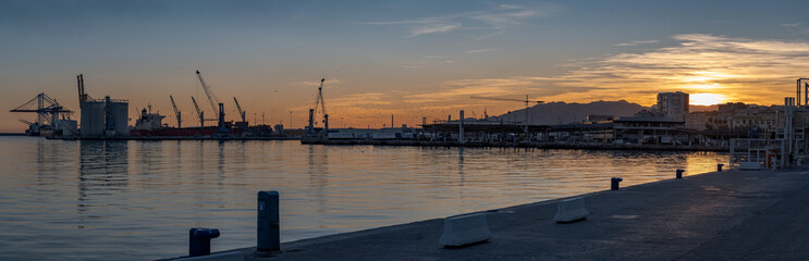 Sunset on the Port of Malaga Spain wide panorama. Docks with cranes silhouette with the sun setting behind the hills in backlight.