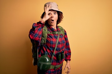 Middle age curly hair hiker woman hiking wearing backpack and water canteen using binoculars doing ok gesture shocked with surprised face, eye looking through fingers. Unbelieving expression.