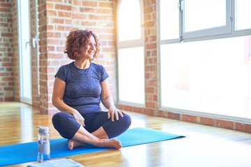 Middle age beautiful sportswoman wearing sportswear sitting on mat practicing yoga at home looking away to side with smile on face, natural expression. Laughing confident.