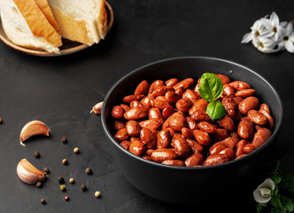 Closeup of a cup with boiled red beans decorated with basil leaves. Healthy vegetarian dish, next to a bowl of spices, bread and a fresh salad. Close-up on a dark vintage background, horizontal view